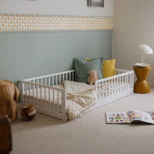 Lit enfant Montessori blanc en bois, modèle Monty 80x160 cm de Woodies, avec barrières de sécurité, coussins colorés, peluche et couverture, dans une chambre décorée de façon douce et moderne.
