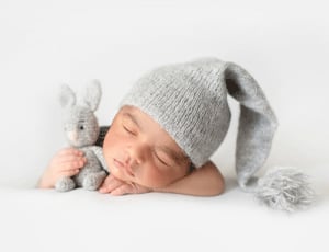 Un bébé endormi portant un bonnet gris avec un pompon et tenant un doudou lapin